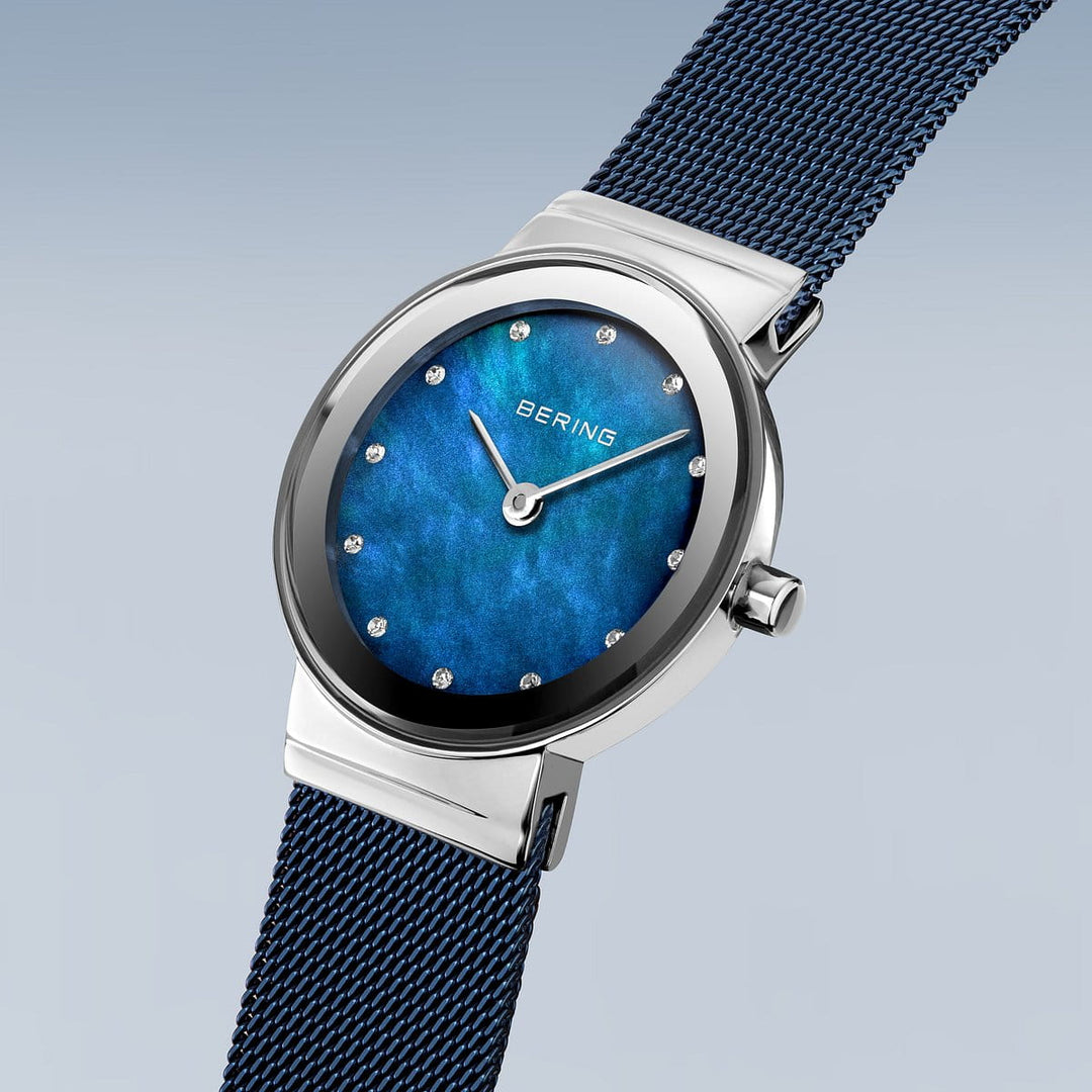 Het Bering 10126-307 dameshorloge heeft een blauwe parelmoer wijzerplaat, zilveren uurmarkeringen en wijzers. Dit quartzhorloge heeft een roestvrijstalen kast, blauwe meshband en saffierglas, afgezet tegen een lichtblauwe naar grijze achtergrond met kleurverloop.