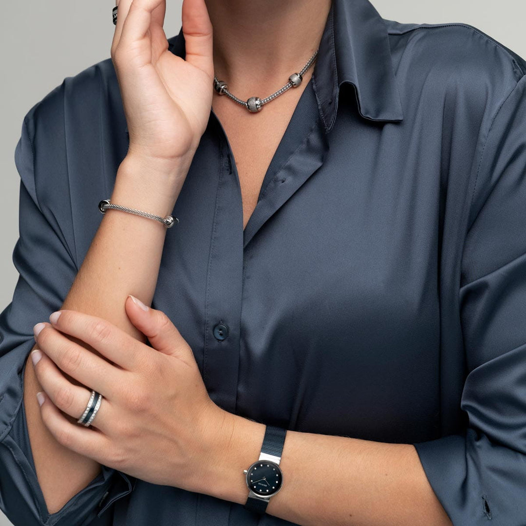 Een persoon toont op elegante wijze sieraden: een ring, armband, ketting en een Bering 10126-307 zwart polshorloge met saffierglas. Ze dragen een zijdezacht donkergrijs overhemd met knopen, met één hand sierlijk omhoog bij hun gezicht terwijl hun haar verborgen blijft.