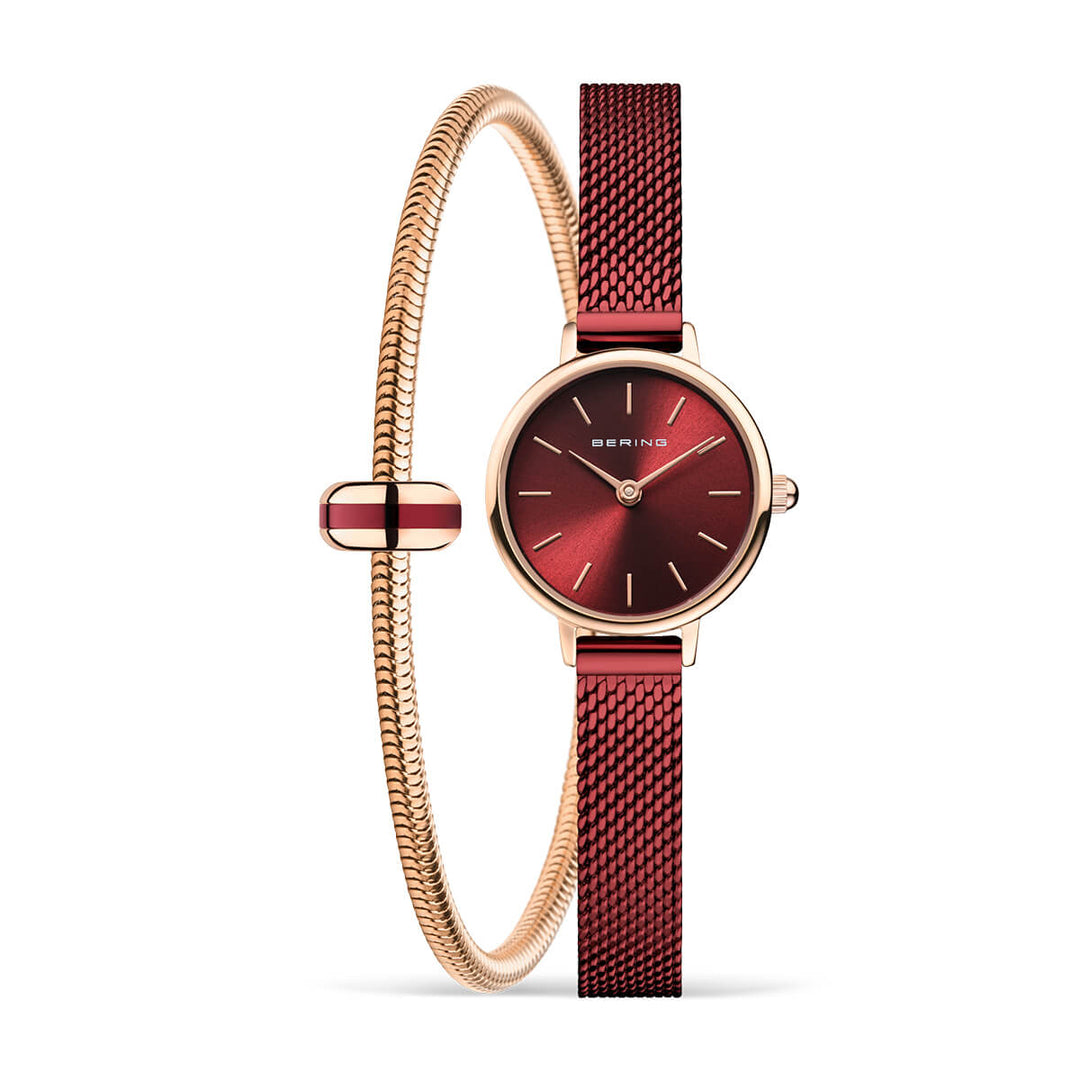 Het Bering 11022-363-Lovely-5-GWP190 horloge heeft een ronde gouden lunette, minimalistische wijzerplaat en rode mesh band met saffierglas. Het wordt vergezeld door een dunne gouden armband met rode accenten. Beide items worden verticaal weergegeven tegen een witte achtergrond.
