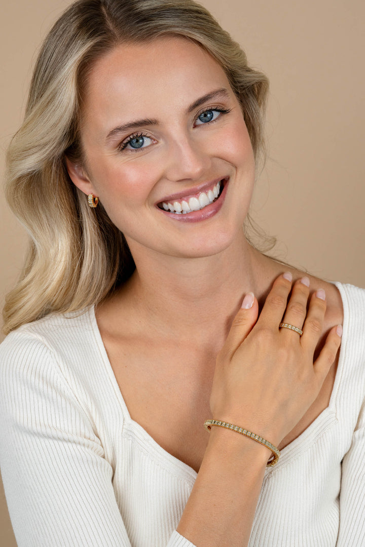 Een lachende blonde vrouw in een wit topje showt de Zinzi Ring Zilver ZIR2686Y van Zinzi, samen met bijpassende tweekleurige gouden sieraden, terwijl ze haar armen over elkaar slaat en naar de camera kijkt tegen een beige achtergrond.