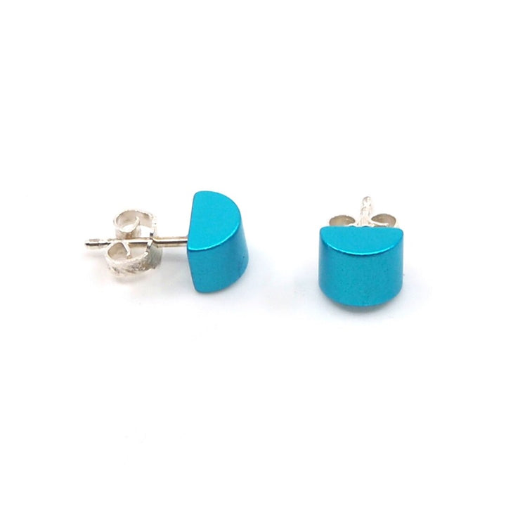 De Otra Cosa Gekleurde kubus oorbellen turquoise bestaat uit twee oorstekers met een halfrond ontwerp in turquoise, geplaatst op een strakke geanodiseerde aluminium achterkant, die een minimalistische en moderne stijl uitstralen tegen een effen witte achtergrond.