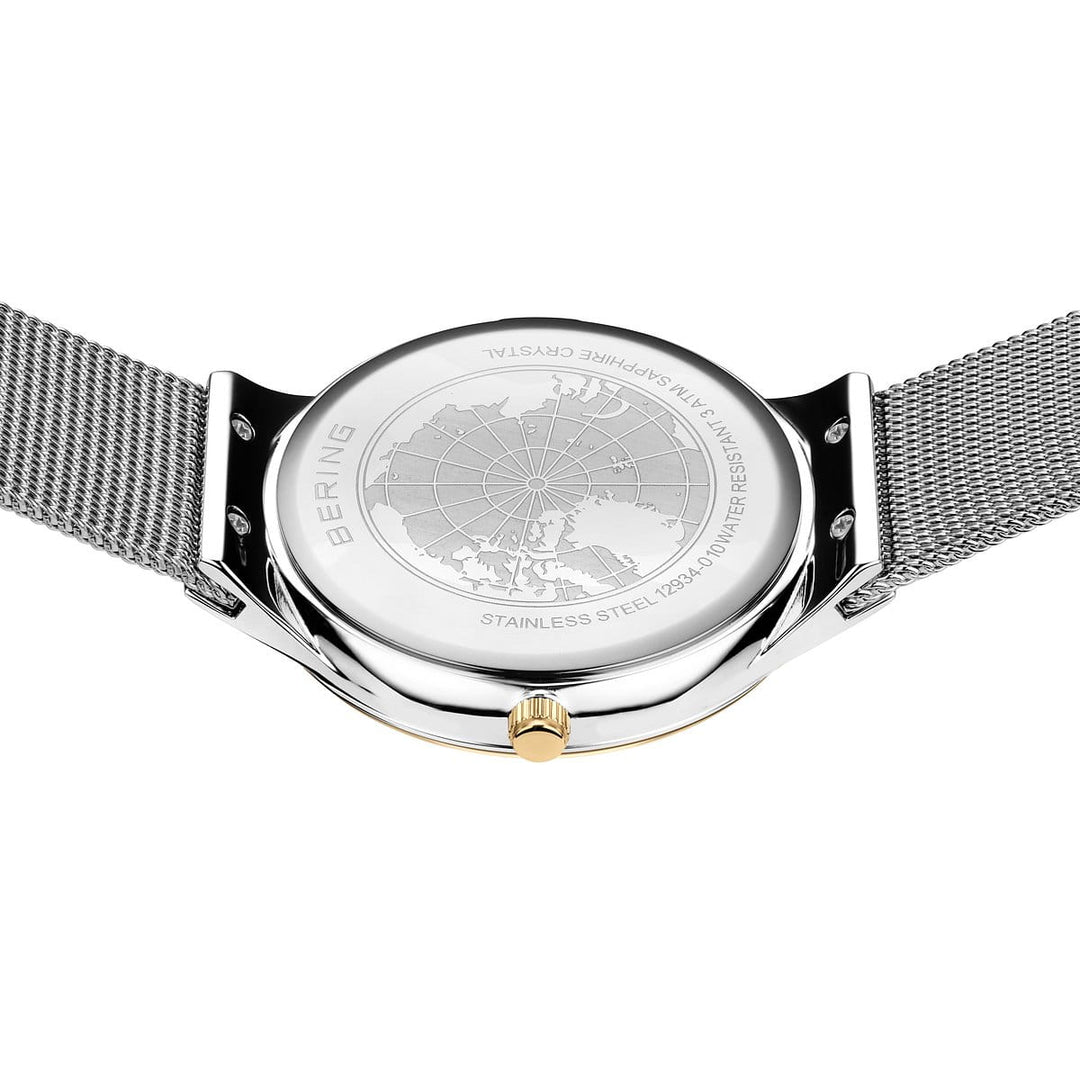 De afbeelding toont de achterkant van een Bering 12934-010 Dameshorloge met een tijdloos design, met een roestvrijstalen kast gegraveerd met een wereldbol en tekst over materialen en waterbestendigheid. Een metalen meshband voegt elegantie toe.