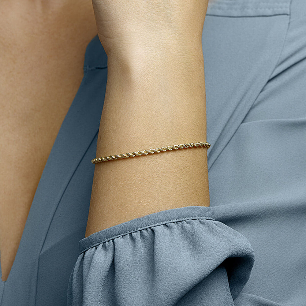 Een close-up laat een persoon zien die de Van der Kooij Collectie armband koord 2.7mm draagt, een eenvoudige en elegante gouden armband, gecombineerd met een lichtblauwe blouse met aangerimpelde mouwen tegen een neutrale achtergrond.