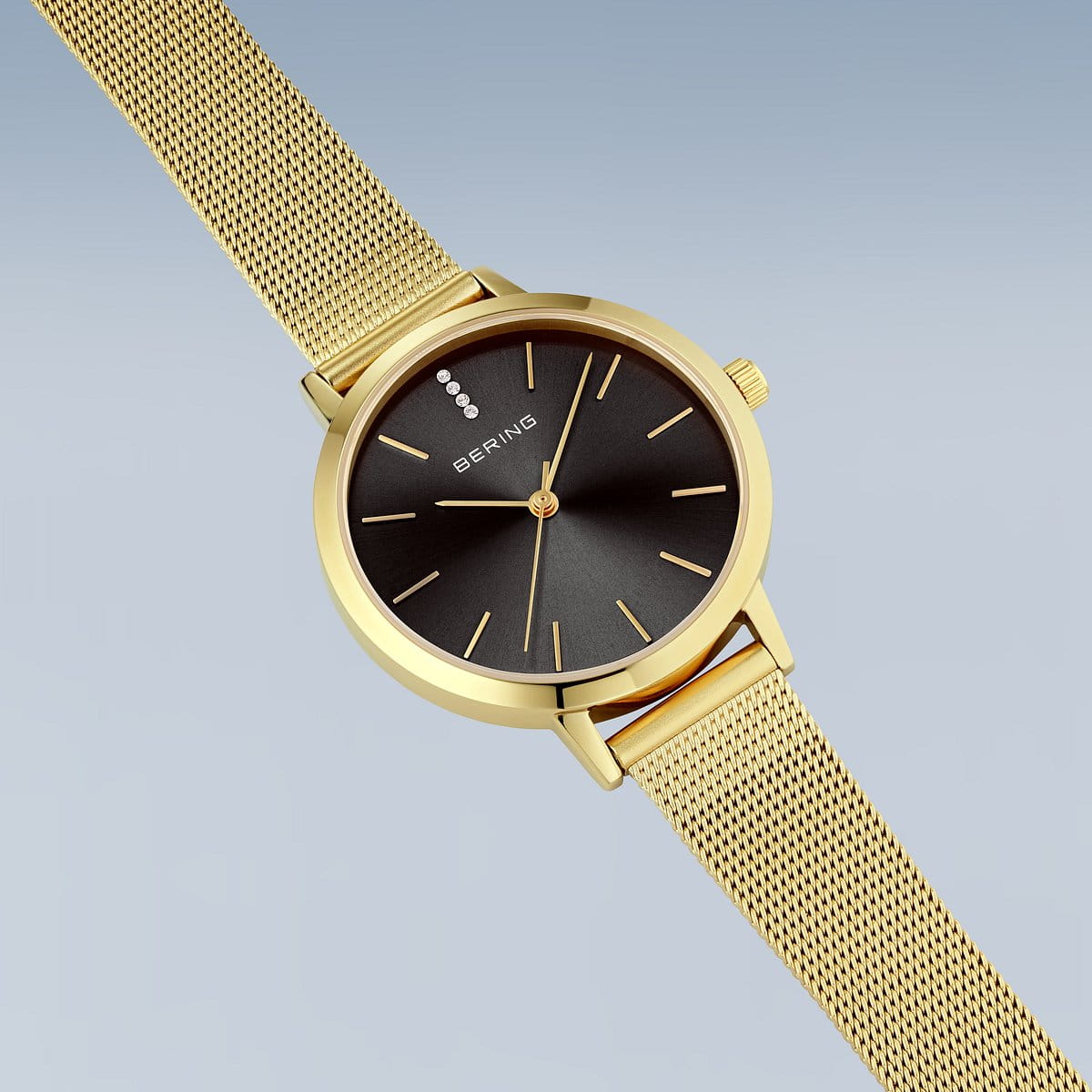 Het Bering dames horloge 13434-332 heeft een zwarte wijzerplaat met gouden minimalistische uurmarkeringen, saffierglas en een goudkleurige Milanees band. BERING staat op de wijzerplaat, gepresenteerd tegen een lichtblauwe achtergrond.