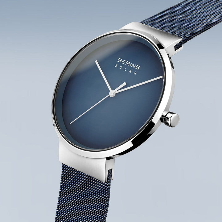 De Bering bering unisex horloge blauwe dial - 14339-307 is een minimalistisch zilveren horloge met een diepblauwe wijzerplaat en dunne zilveren wijzers, aangevuld met een mesh band. Het straalt moderne stijl uit tegen een lichtblauwe gradiëntachtergrond, waardoor het een elegante accessoire is.