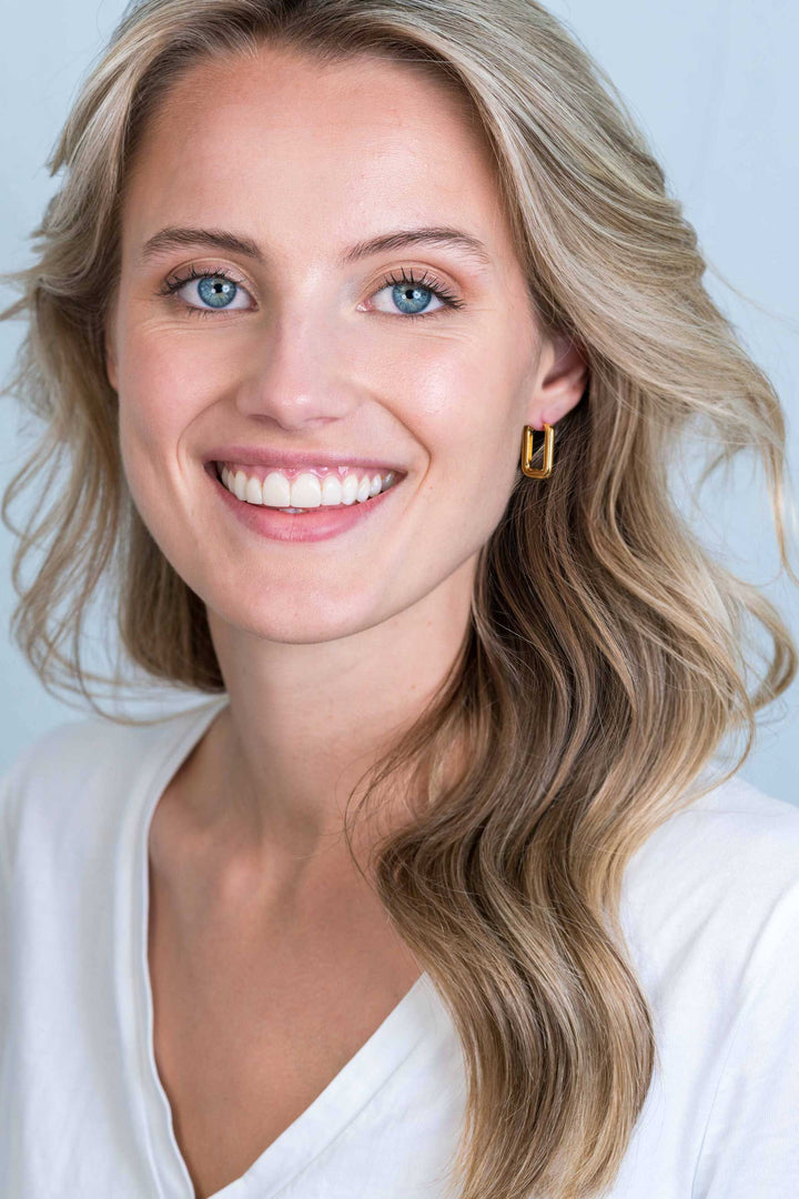 Een jonge vrouw met lang, golvend blond haar en blauwe ogen lacht naar de camera, draagt een wit topje en een enkele Zinzi Oorbellen Gold Plated Zio2221 G van Zinzi. De achtergrond is licht en effen.
