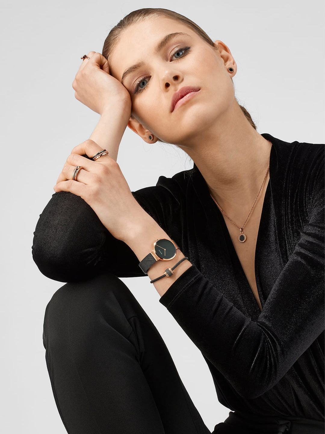 Een vrouw in een zwarte outfit zit met haar elleboog op haar knie, en toont een verbluffend Bering dames horloge 14531-166 met een roestvrijstalen kast. Haar look wordt aangevuld met verschillende sieraden, waaronder een ketting en verschillende ringen. Ze behoudt een neutrale uitdrukking tegen een effen grijze achtergrond.