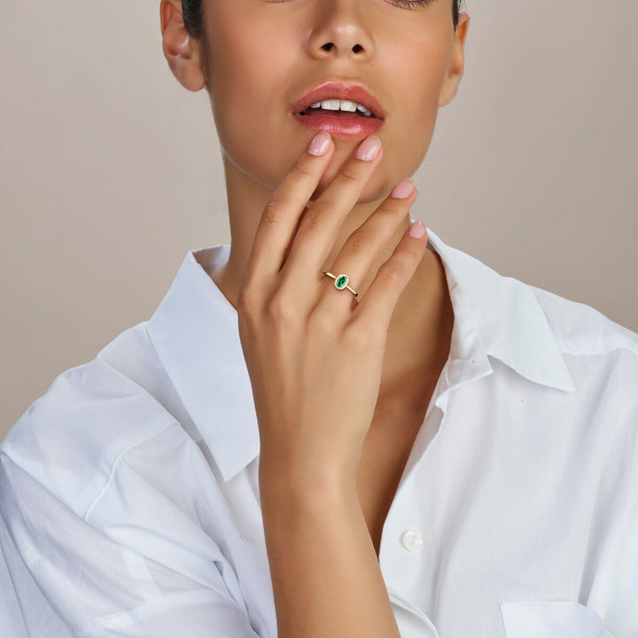 Een vrouw in een wit overhemd raakt haar gezicht aan, waardoor de Van der Kooij Collectie ring lab grown smaragd en diamant 0.075ct H SI halo 14K geelgoud aan haar vinger wordt benadrukt tegen een neutrale achtergrond.