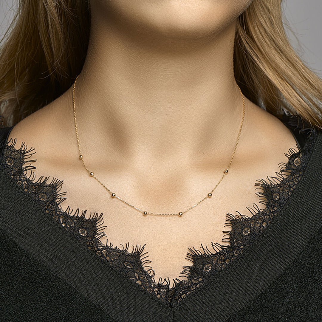 Een vrouw draagt een Van der Kooij Collectie collier anker en bolletjes 1,0 mm 42-44-46 cm 14K geelgoud, de delicate gouden kralen verspreid langs de ketting, gecombineerd met een zwart kanten topje. Alleen haar nek en bovenborst zijn zichtbaar.