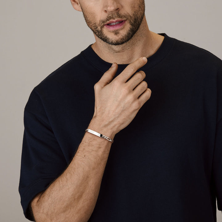 Een man met een getrimde baard in een marineblauw t-shirt raakt zijn nek aan, waardoor de Van der Kooij Collectie graveerarmband figaro plaat 6,0 mm zilver wit om zijn pols subtiel wordt benadrukt tegen een neutrale achtergrond.