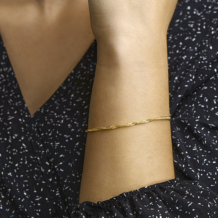 Een close-up van de arm van een persoon die de Van der Kooij Collectie armband singapore 2,3 mm 14K geelgoud draagt, gecombineerd met een zwart topje met witte spikkelpatronen.