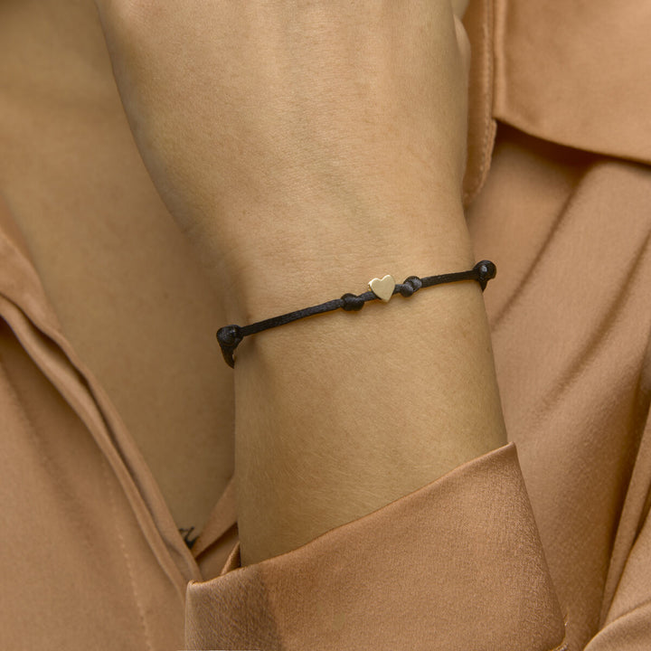 Een persoon in een perzikkleurige blouse draagt de Van der Kooij Collectie armband satijn hart 14K goud met rubber/nylon/leer om zijn pols - een zwart gevlochten bandje met een gouden hartje - terwijl hij zachtjes zijn gezicht aanraakt.