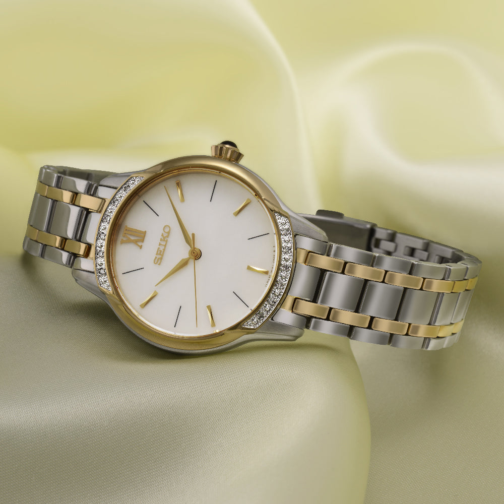 Het Seiko dameshorloge SRZ544P1 heeft een zilveren en gouden metalen band, een witte wijzerplaat met gouden markeringen en wijzers, omlijst door een roestvrijstalen kast. Fonkelende stenen sieren de lunette, allemaal afgezet tegen een zachtgele stoffen achtergrond voor een elegante touch.