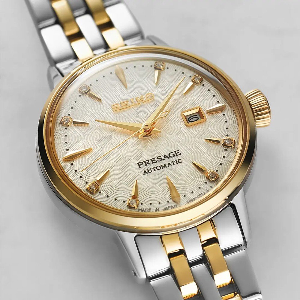 De Seiko Presage dames horloge SRE010J1 van Seiko met automatisch uurwerk, goud-zilveren band, crème textuur wijzerplaat, gouden markeringen en datum op 3 uur is te zien tegen een lichte achtergrond.