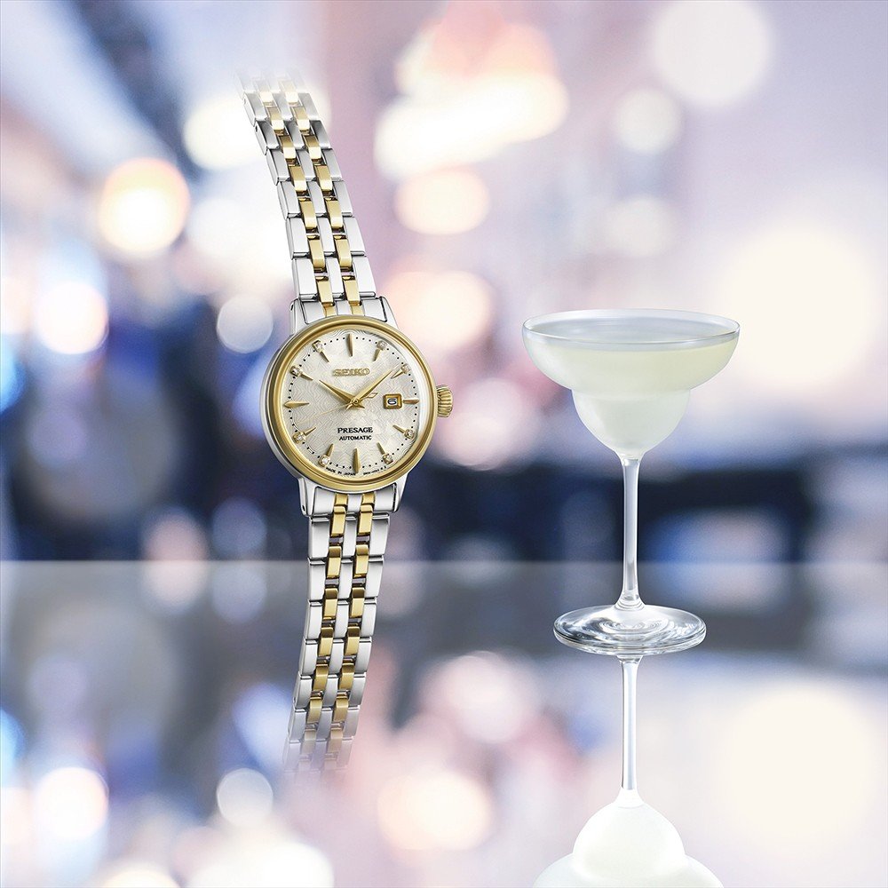 De Seiko Presage dames horloge SRE010J1 staat rechtop naast een coupeglas met een lichte cocktail, beide gespiegeld op een glanzend oppervlak met een zacht onscherpe achtergrond.
