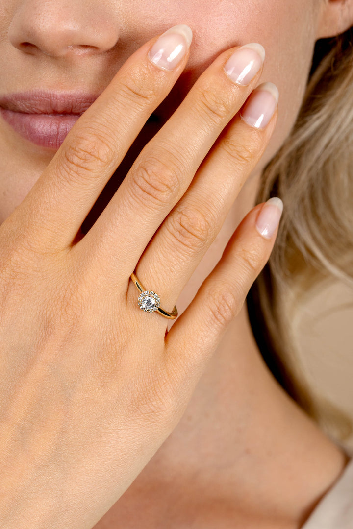 Een close-up van de verzorgde hand van een vrouw die de Zinzi Ring Gold Plated ZIR2697Y van Zinzi draagt, met een ronde, sprankelende witte zirkonia omringd door kleinere stenen, dicht bij haar gezicht.