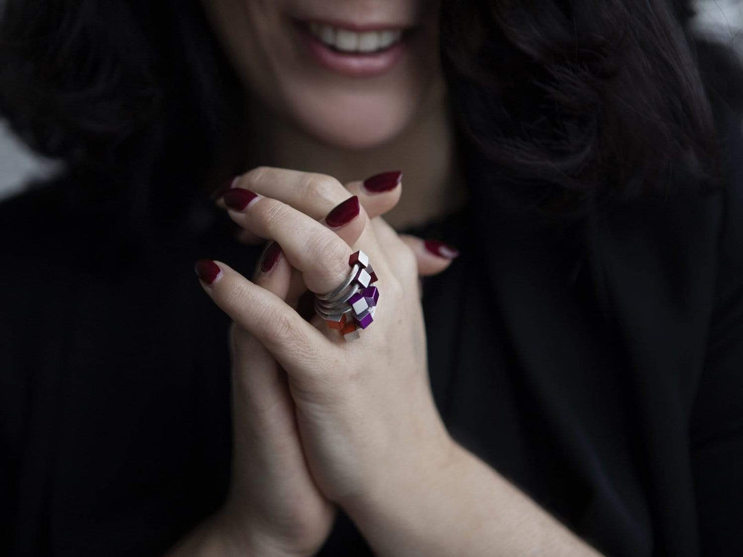 Een persoon met donker haar en bordeauxrode nagellak vouwt zijn handen en toont CLIC Creations Ring Kubusjes Rood R4R. Gemaakt van chirurgisch edelstaal in paarse en zilveren geometrische vormen, benadrukken de handgemaakt in Nederland ringen een zachte glimlach en complementeren ze perfect een donkere outfit.