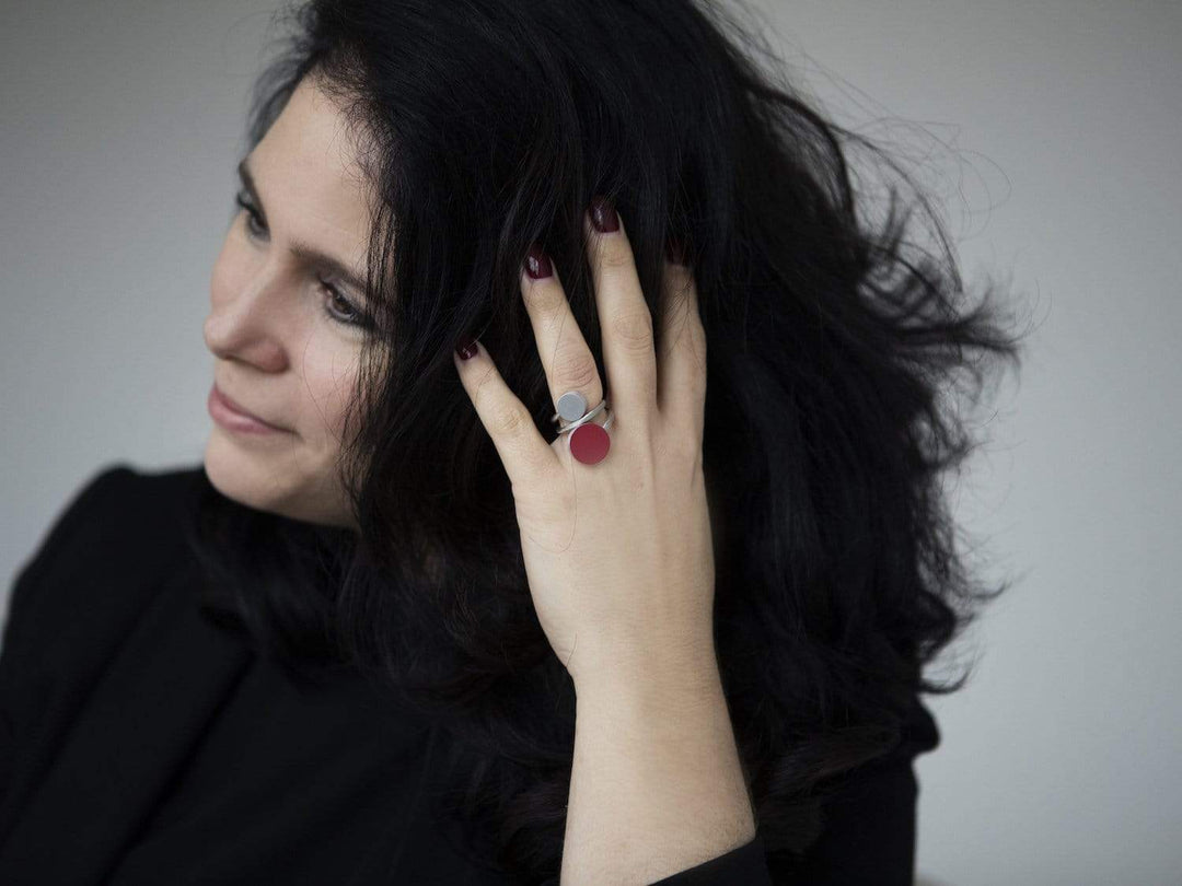 Een persoon met lang donker haar raakt zijn hoofd aan en toont de CLIC Creations Ring Twee ronde vormen Goud|Zand R5G met unieke rode en grijze cirkels. Met kastanjebruine nagels tegen een neutrale achtergrond benadrukken ze de handgemaakte in Nederland allure van dit accessoire.