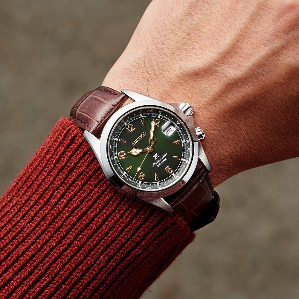 Een persoon draagt een Seiko Prospex SPB121J1, met een groene wijzerplaat, zilveren uurmarkeringen en een bruine leren band. Dit automatische horloge is waterbestendig tot 20ATM. Als aanvulling op het horloge dragen ze een roodbruine geribbelde trui.