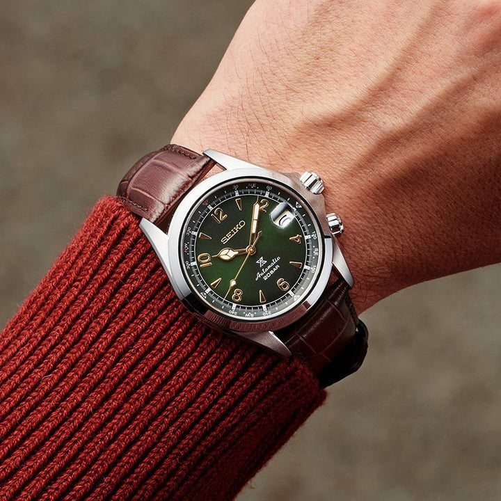 Een persoon draagt een Seiko Prospex SPB121J1, met een groene wijzerplaat, zilveren uurmarkeringen en een bruine leren band. Dit automatische horloge is waterbestendig tot 20ATM. Als aanvulling op het horloge dragen ze een roodbruine geribbelde trui.