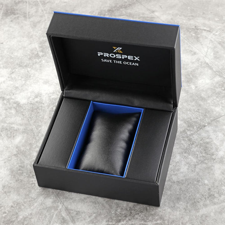 Een open Seiko Prospex SRPE35K1 doos met een blauwe binnenkant rust op een grijs oppervlak. Binnenin zit een zwarte kusseninzet, waarschijnlijk voor een horloge, en op het deksel staat PROSPEX SAVE THE OCEAN.