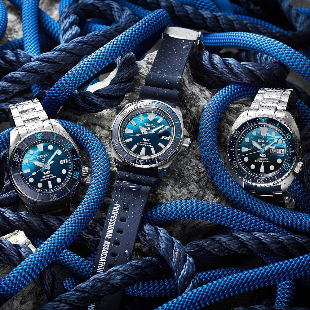Drie Seiko Prospex duikhorloges, elk met blauwe wijzerplaten en roestvrijstalen kasten, hebben natte blauwe touwen. De linker- en rechterhorloges hebben metalen armbanden, terwijl het middelste horloge een rubberen band heeft. De gestructureerde achtergrond accentueert het nautische thema.