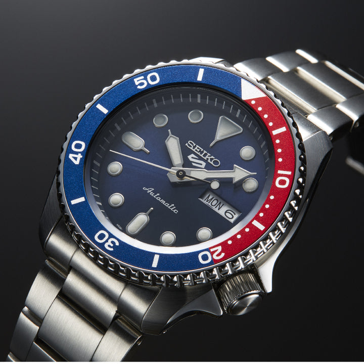 Een close-up van de Seiko 5 Sports heren horloge SRPD53K1 van Seiko, met een metalen band, blauwe en rode lunette, donkerblauwe wijzerplaat met lichtgevende markeringen en een automatisch uurwerk met dag-datum aanduiding (MON/6) op een zwarte achtergrond.