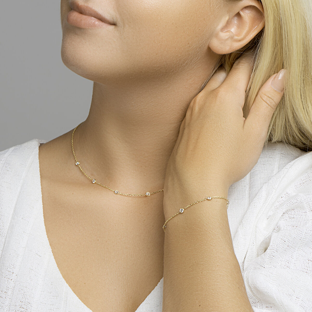 Een vrouw in een wit getextureerd topje met V-hals raakt zachtjes haar nek aan en draagt de Van der Kooij Collectie gouden dames ketting zirkonia zilver verguld (geel) en bijpassende armband - elegantie in elk detail.