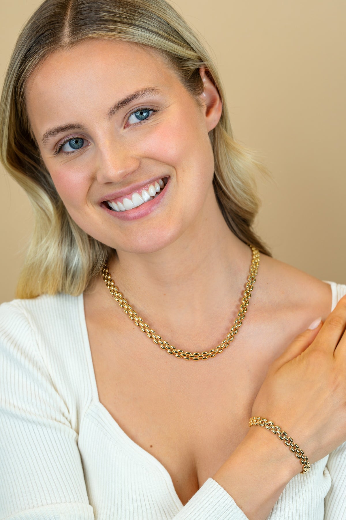 Een lachende blonde vrouw in een wit topje modelleert de Zinzi Armband Gold Plated ZIA2764G tegen een beige achtergrond.