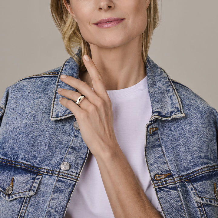 Een vrouw in een spijkerjasje en wit t-shirt raakt haar hals aan en pronkt met een Van der Kooij Collectie ring zilver gerhodineerd aan haar vinger. Haar gezicht is deels zichtbaar met een lichte glimlach.