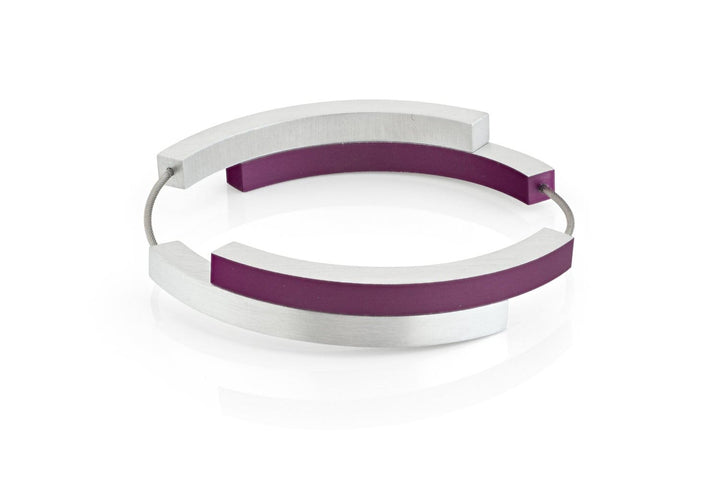 De Armband Vier bogen Paars A32P van CLIC Creations is een moderne armband gemaakt in Nederland. Het heeft twee gebogen zilveren metalen bogen en twee paarse bogen verbonden door een dunne, flexibele draad met een magnetische sluiting, die een cirkelvormig ontwerp vormen op een witte achtergrond.
