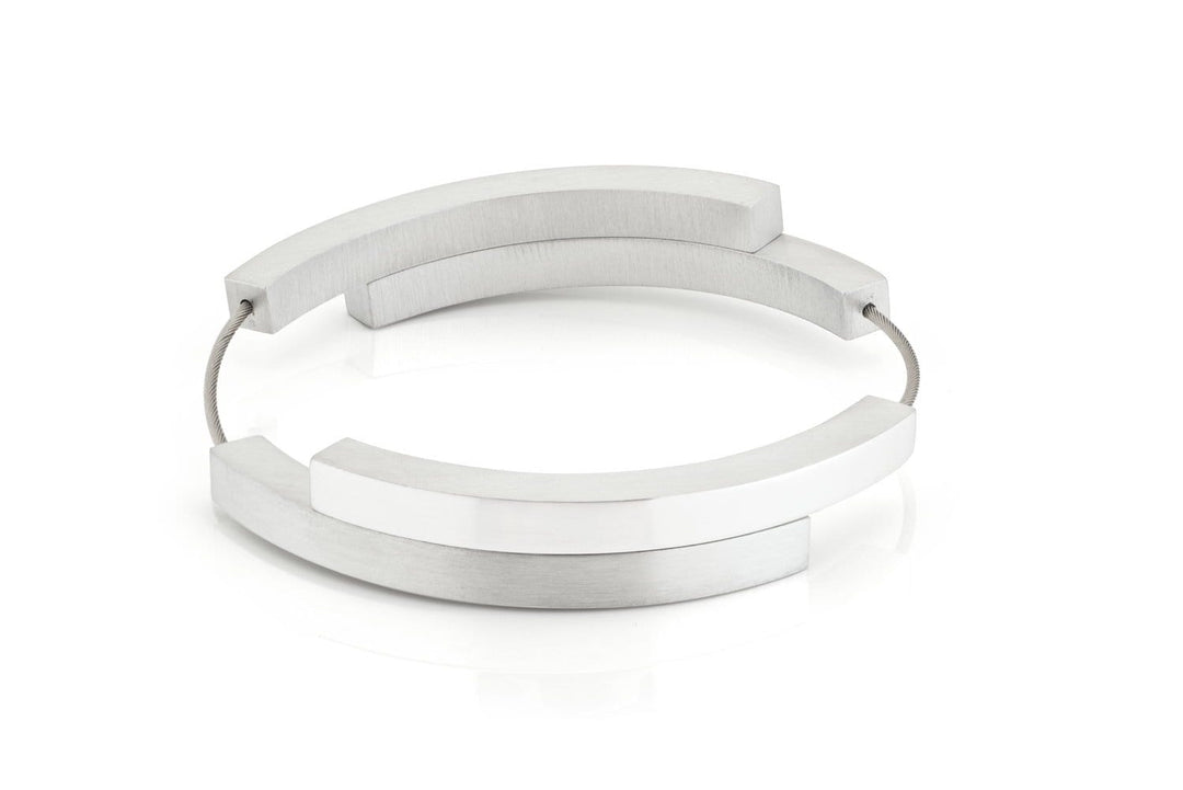 De Armband Vier bogen mat|glans A32 van CLIC Creations is een moderne, minimalistische armband met overlappende zilveren bogen die met elkaar verbonden zijn door een dunne draad en heeft een magnetische sluiting. Dit geometrische stuk is handgemaakt in Nederland en wordt tentoongesteld tegen een effen witte achtergrond.