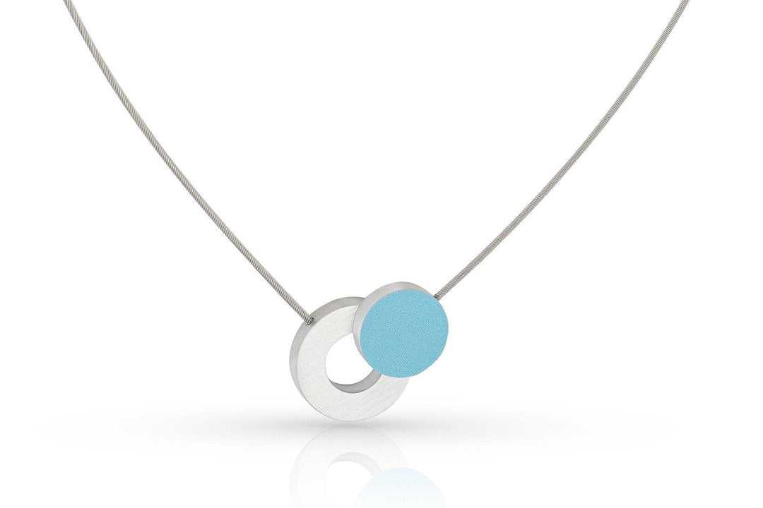 De Collier Gekleurd rondje en een ring C265B van CLIC Creations heeft een minimalistisch ontwerp met een dunne zilveren ketting, twee in elkaar grijpende hangers - een zilveren open cirkel en een effen hemelsblauwe cirkel - en een magnetische sluiting. Dit handgemaakte stuk uit Nederland valt op tegen de witte achtergrond.