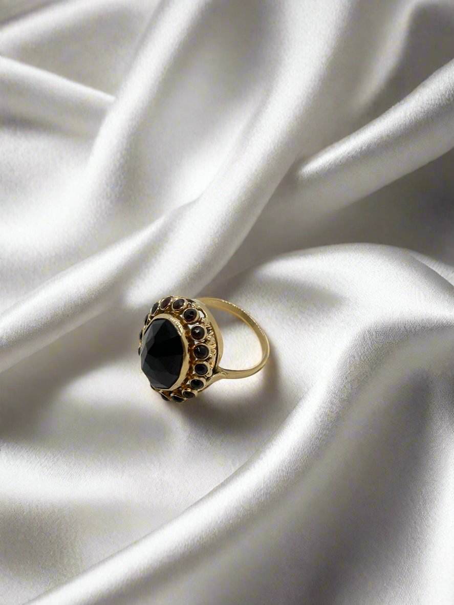 Een vintage gouden ring 14k ovale granaat 5,9g van Juwelier van der Kooij, met een grote zwarte edelsteen en kleinere donkere stenen, rust op glad wit satijn. Elegante plooien versterken de schoonheid uit onze Vintage Collectie.