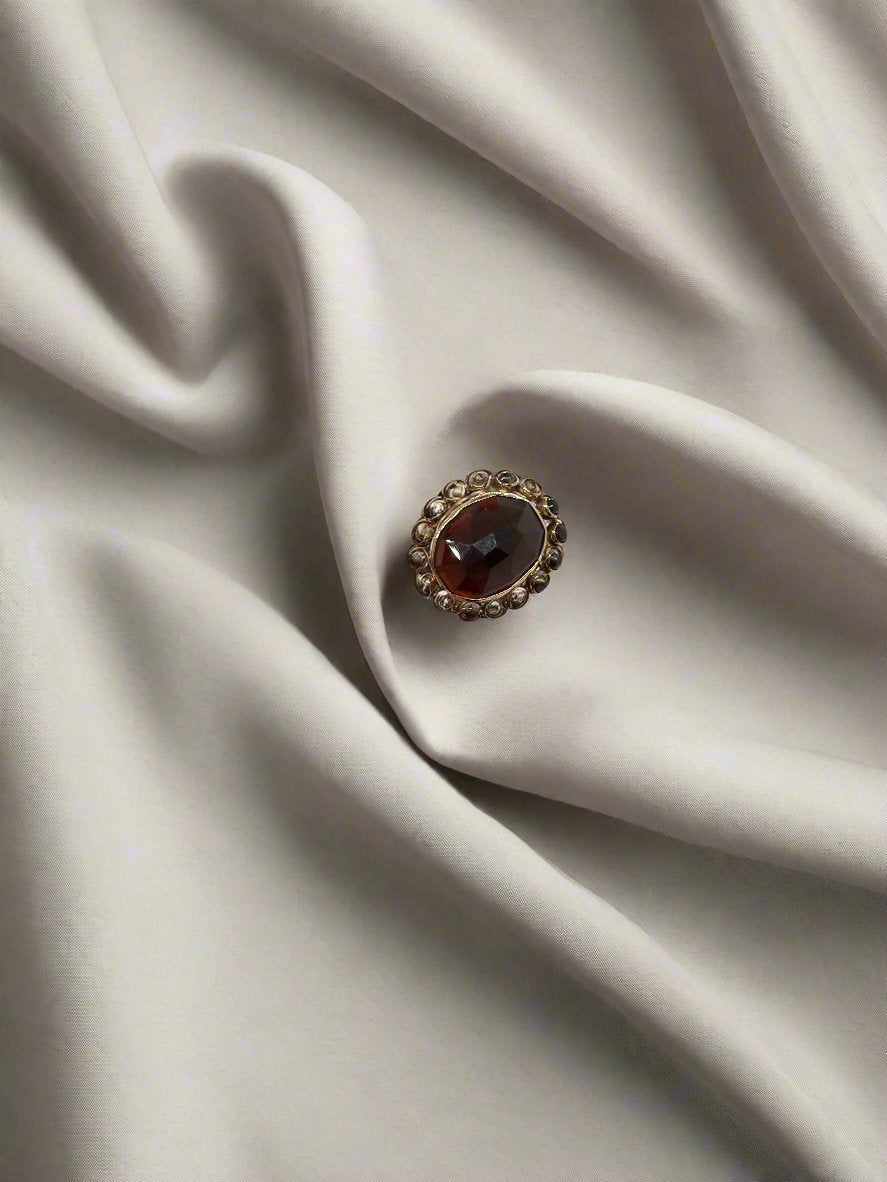 Een vintage gouden ring van 14 karaat van Juwelier van der Kooij, met een ovale granaat edelsteen van 5,9 gram, is gezet op een gladde, lichtgrijze stof waarbij de vouwen zachte schaduwen werpen over dit tijdloze sieraad.