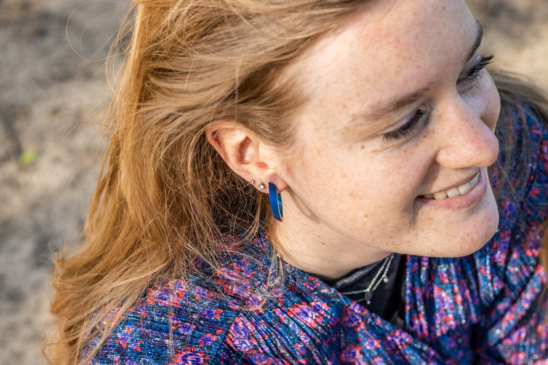 Een persoon met lang blond haar en blauwe oorbellen van chirurgisch edelstaal glimlacht terwijl hij opzij kijkt, gekleed in een gebloemd blauw en paars kledingstuk. Hun ovale vorm creolen, Ovale creolen O21 van CLIC Creations, dansen in de wind tegen een zanderige achtergrond.