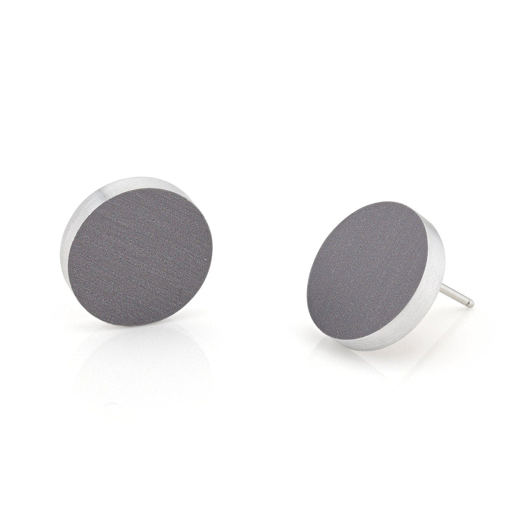 De Oorsteker rond O53AG oorbellen van CLIC Creations zijn handgemaakt in Nederland en hebben een glad donkergrijs oppervlak met zilveren randen. Eén oorbel staat rechtop, terwijl de andere gekanteld is om de pin te onthullen tegen een schone, reflecterende witte achtergrond.