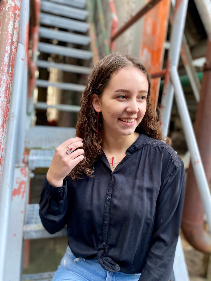 Een jonge vrouw met krullend haar, in een zwarte blouse en blauwe jeans, zit op een metalen trap. Ze glimlacht terwijl ze opzij kijkt en de Collier Eenvoudige staafjes Oranje C116O van CLIC Creations om haar nek aanraakt. De achtergrond toont versleten, veelkleurige leuningen.