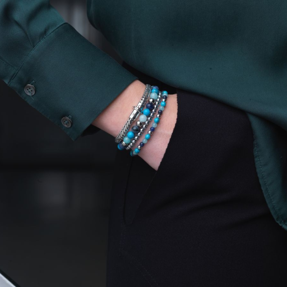 Een persoon in een donkergroen shirt met lange mouwen en een zwarte broek heeft zijn hand in zijn zak en toont gestapelde blauwe en zilveren armbanden, waaronder de Rebel & Rose Blue Summer Vibes - 6mm Kralen Armband Dames.