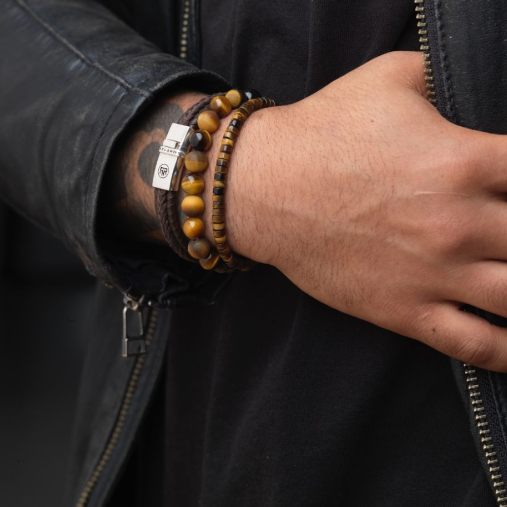 Een persoon met een zwart leren jack en shirt toont zijn pols versierd met meerdere aarde-tone heren armbanden, waaronder de Rebel & Rose Tiger Lily - 8mm Kralen Armband Heren met een klein metalen label.