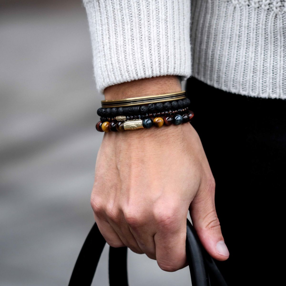 Een hand die de Rebel & Rose Black Moon - All-Black 8mm heren kralen armband draagt, houdt een zwart handvat vast. Andere kralen armbanden heren en een witte gebreide trui mouw zijn ook zichtbaar.