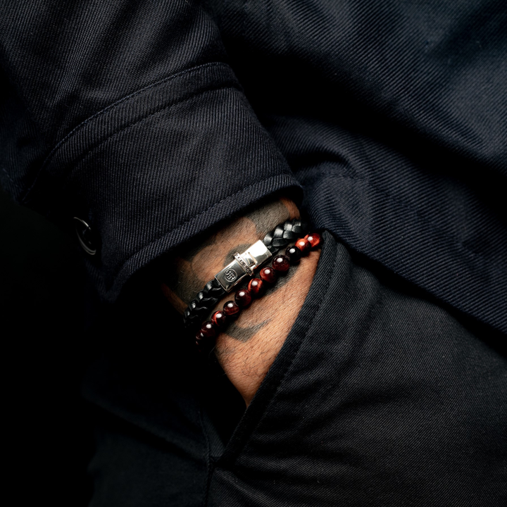 Een persoon in een donker shirt met lange mouwen draagt de Rebel & Rose Small Braided Raw Black leren heren armband met een zilveren sluiting, samen met drie kralenarmbanden-twee zwarte en een roodbruine. Een stijlvol cadeau-idee voor hem.