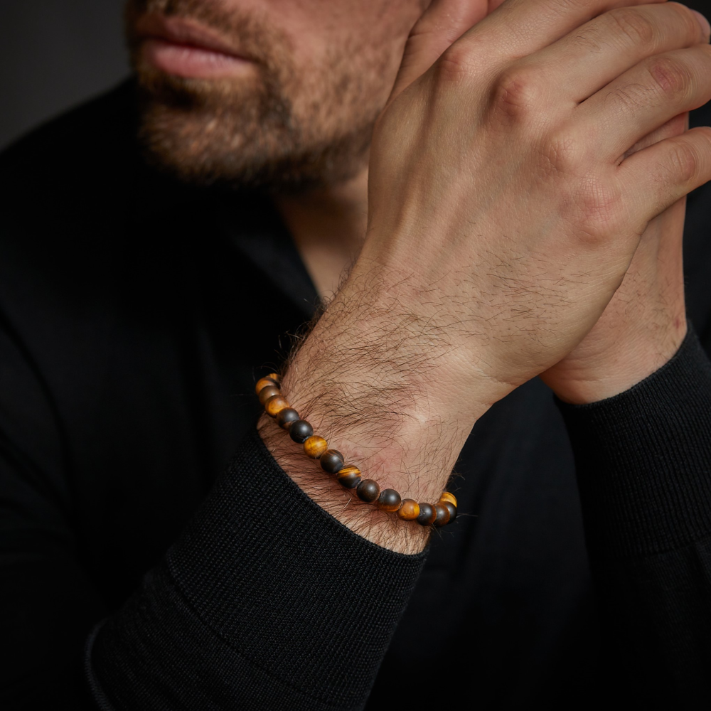 Een bebaarde man in een zwart shirt met lange mouwen omklemt zijn handen en toont de Rebel & Rose Tiger Lily - 8mm Kralen Armband Heren, een stijlvolle herenarmband met bruine en zwarte kralen om zijn pols.