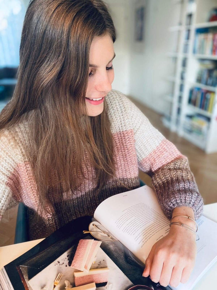 Een persoon met lang bruin haar in een gestreepte trui leest een boek aan een tafel met wafer cookies, een vork en de Thinking of You 🌸 armband basic van CLIC Creations. Op de achtergrond zijn wazige boekenplanken te zien.