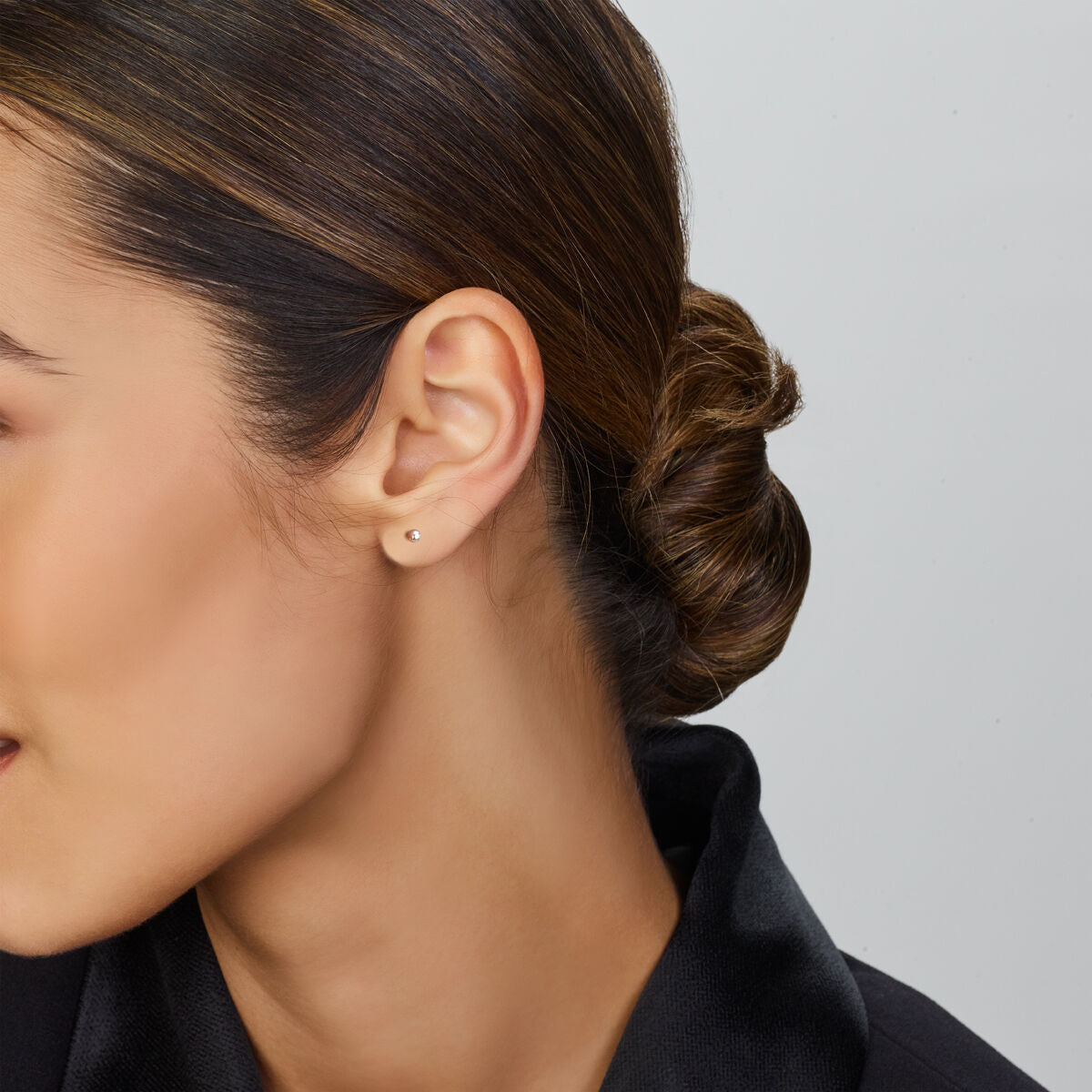 In profiel wordt een vrouw met bruin haar in een laag knotje getoond, gekleed in Van der Kooij Collectie oorknoppen bolletje zilver gerhodineerd en een overhemd met zwarte kraag tegen een effen lichte achtergrond.