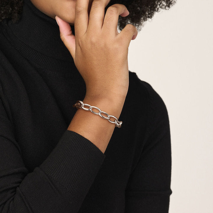 Een persoon in een zwarte coltrui toucheert zijn gezicht en benadrukt de Van der Kooij Collectie armband anker 10 mm 19 cm zilver gerhodineerd om zijn pols tegen een neutrale achtergrond.