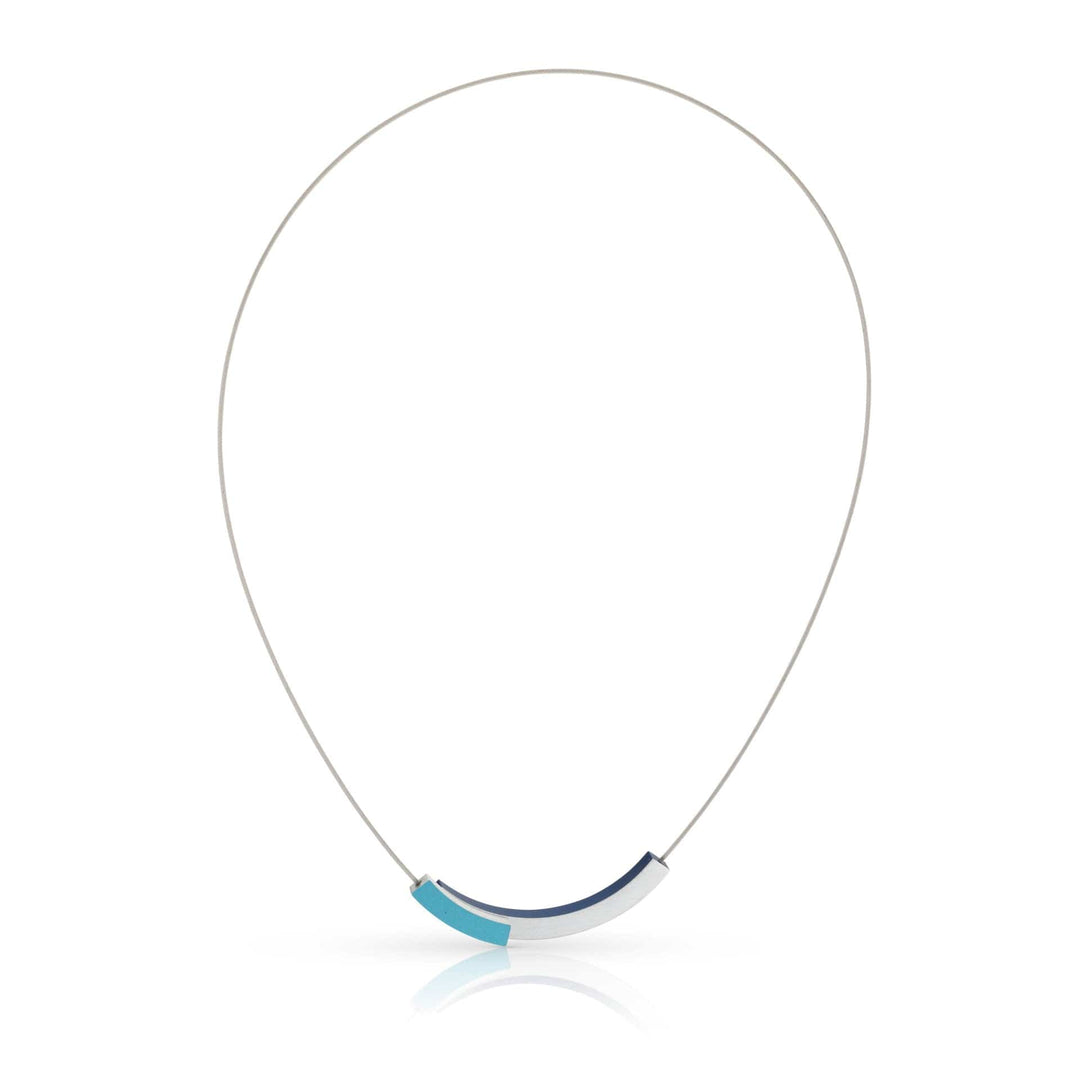 De CLIC Creations Collier twee boogjes met kleur C271B is een minimalistische edelstalen ketting met een strakke hanger van blauw gradiënt gebogen segmenten gecentreerd op een dunne draad. Handgemaakt in Nederland, voorzien van een magnetische sluiting en werpt subtiele schaduwen op een ongerepte witte achtergrond.
