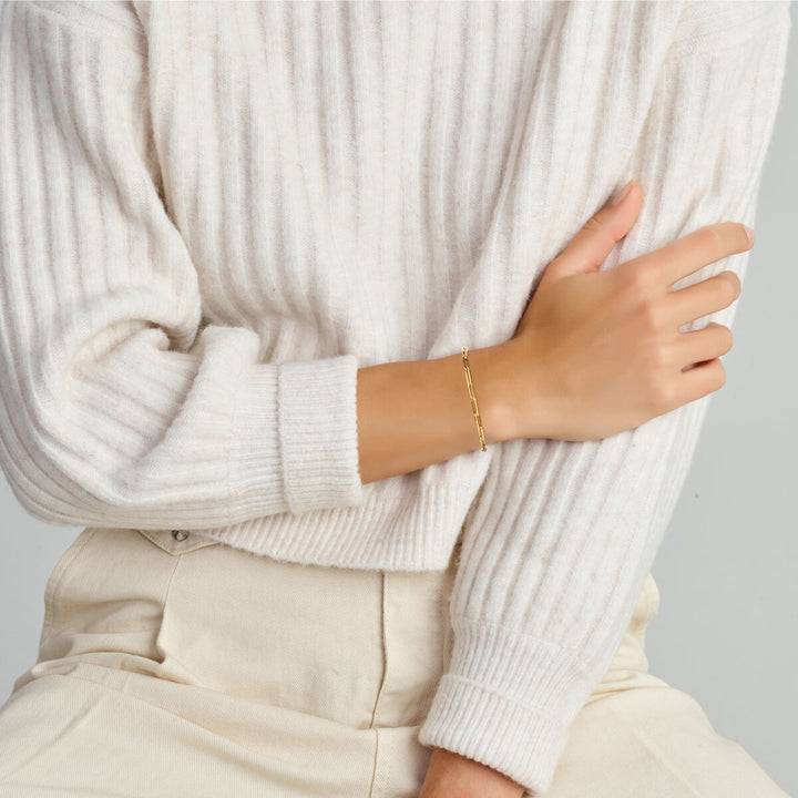 Een persoon in een crèmekleurige geribbelde trui en lichte broek zit met één arm gekruist en toont de Van der Kooij Collectie armband paperclip vierkante buis 3,0 mm 18 cm met groot springslot in 14K geelgoud.