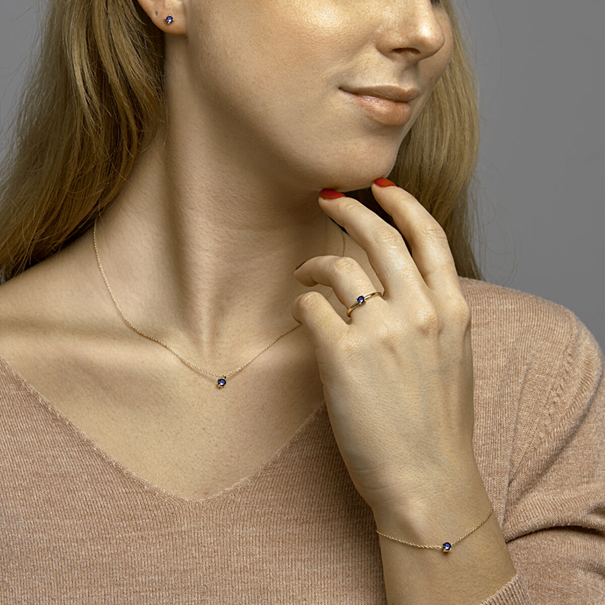 Een vrouw met lang blond haar modelleert de Van der Kooij Collectie Gouden dames ketting saffier 14K-een delicate beige V-hals trui gecombineerd met bijpassende armband, ring en oorbellen, elk met een kleine blauwe saffier.