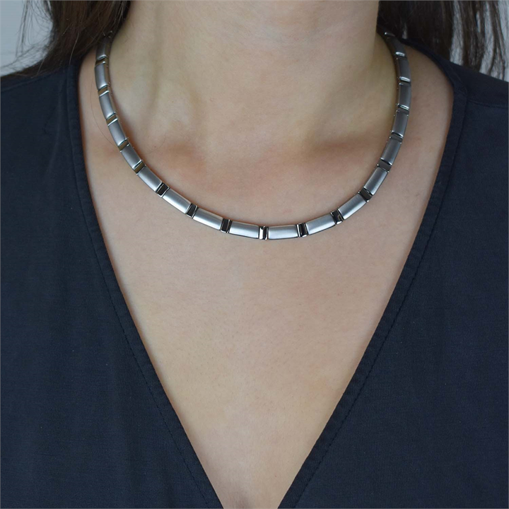 Een close-up toont iemand die de Boccia Titanium Collier 0845-01 draagt, een zilveren, matte ketting met een gesegmenteerd ontwerp. Het individu is gekleed in een donkere top met V-hals tegen een onopvallende achtergrond.